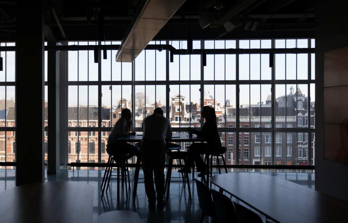 Trois personnes sont assises autour d'une table, image en contre-jour donc très sombre. Le fond est une grande vitre donnant sur le haut d'un immeuble en brique rouge