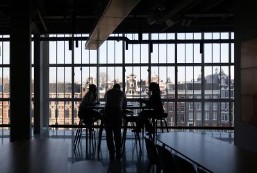 Trois personnes sont assises autour d'une table, image en contre-jour donc très sombre. Le fond est une grande vitre donnant sur le haut d'un immeuble en brique rouge