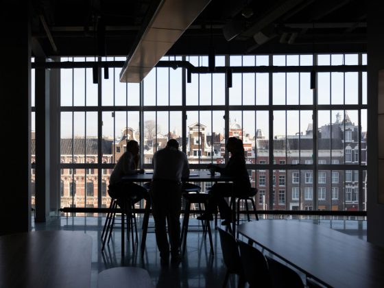 Trois personnes sont assises autour d'une table, image en contre-jour donc très sombre. Le fond est une grande vitre donnant sur le haut d'un immeuble en brique rouge